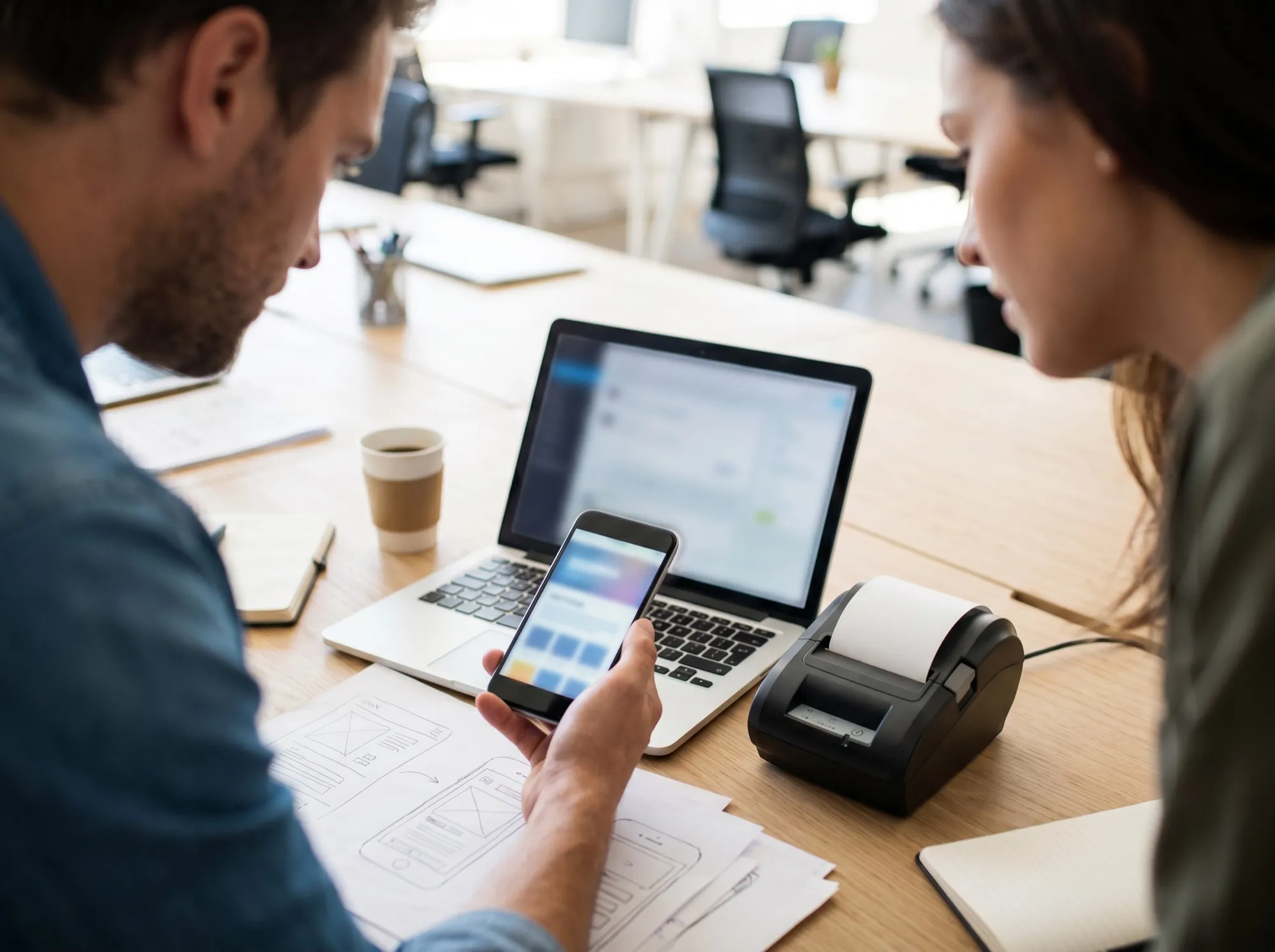 Zwei Personen stimmen eine individuelle Kassen-App ab — Handy, Laptop und Bondrucker am Schreibtisch